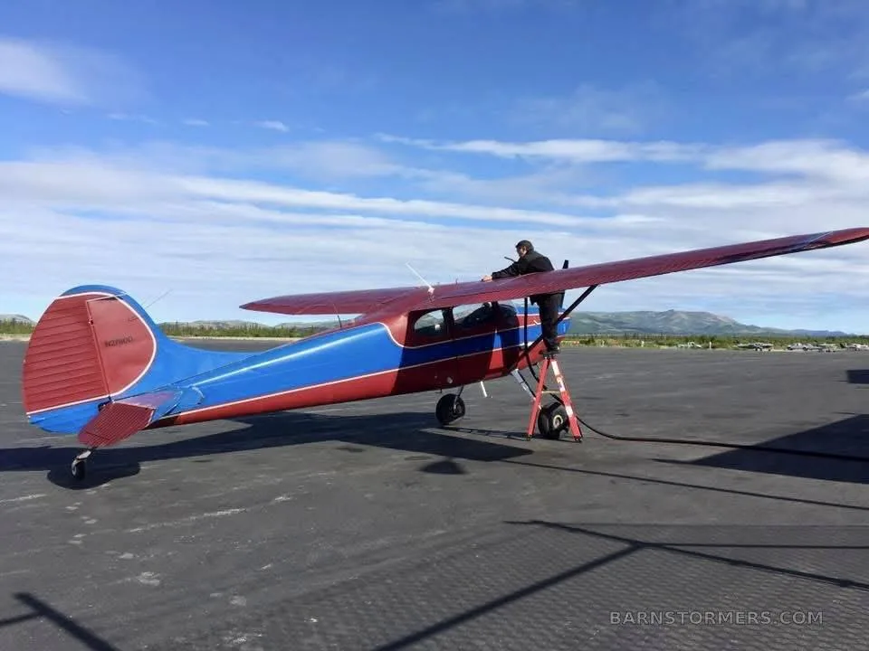 A red and blue high-wing aircraft with a tailwheel, being refueled on an asphalt tarmac, with a person on a ladder.