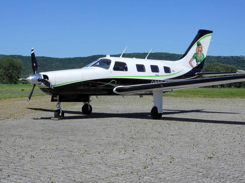 A white and black Piper M-Class aircraft with green accents and a pin-up girl on the tail, parked on a paved surface.