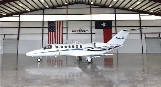 A white Cessna Citation business jet with blue stripes, tail number N525TK, parked in a hangar with US and Texas flags.
