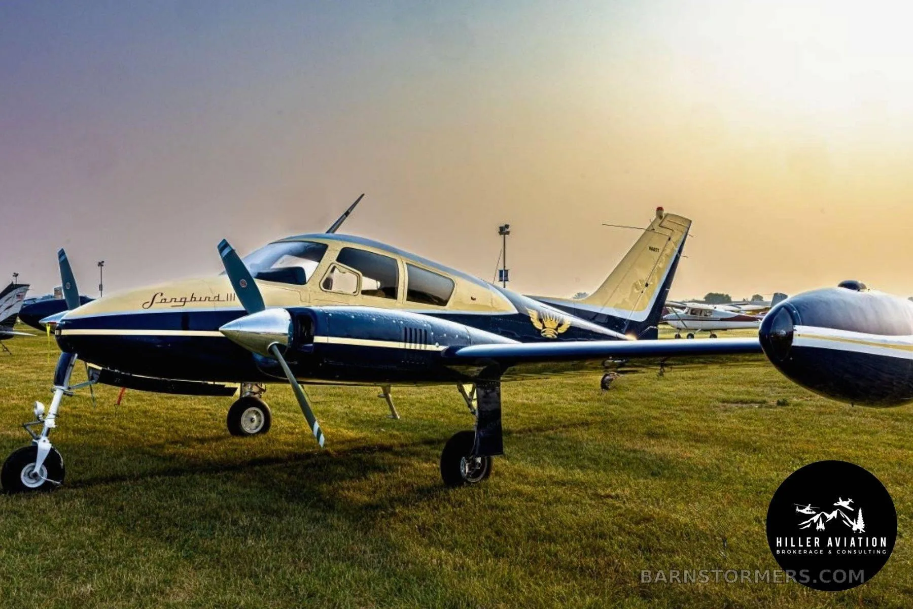 A blue and gold twin-engine aircraft, "Longbird III", is parked on a grassy field at sunset.