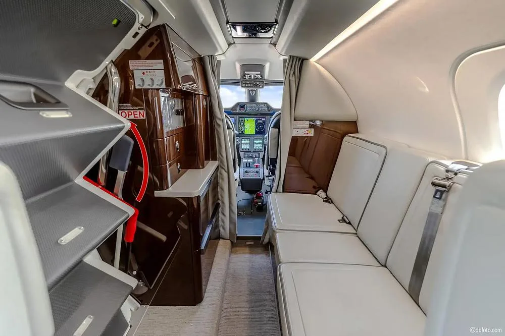 A view from the rear of the private jet cabin, showing a side bench seat, storage compartments, and a glimpse into the cockpit.