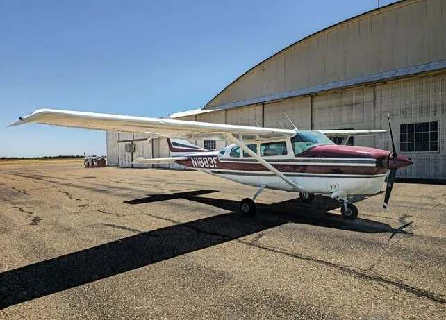 A white and maroon Cessna 210 Centurion aircraft with registration N1883F parked on an airfield next to a large hangar.