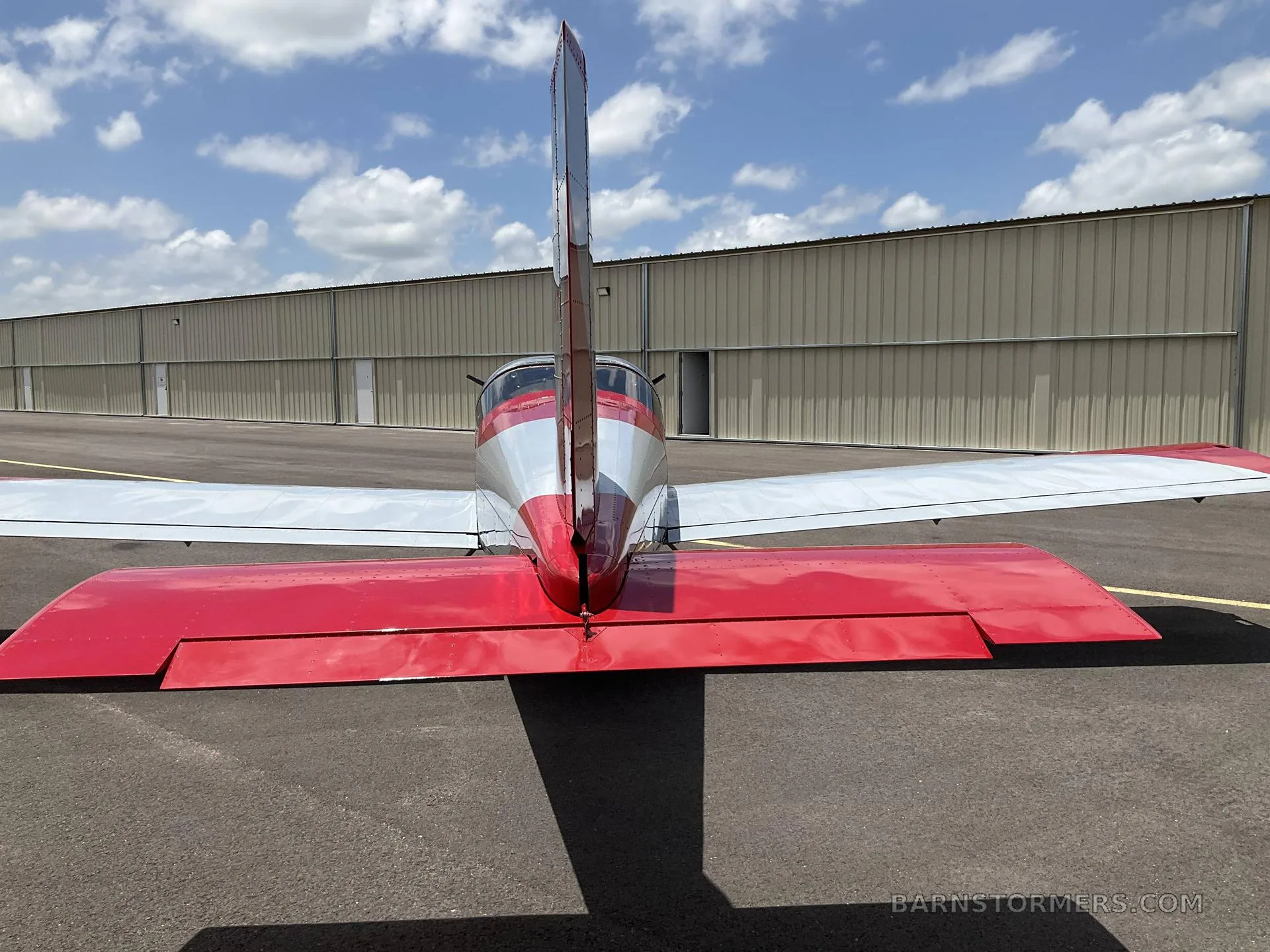 A rear view of a red and grey single-engine aircraft, showing its tail and wings, parked on an asphalt surface in front of a hangar.