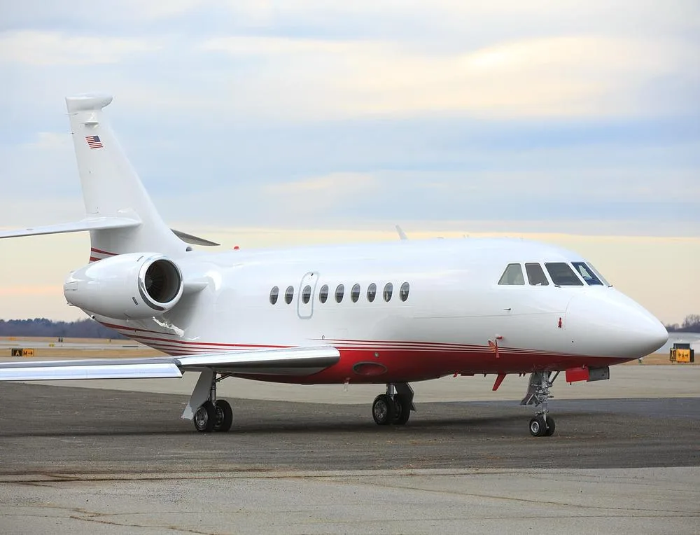 A white and red Dassault Falcon 900 private jet parked on an airport tarmac under a cloudy sky.