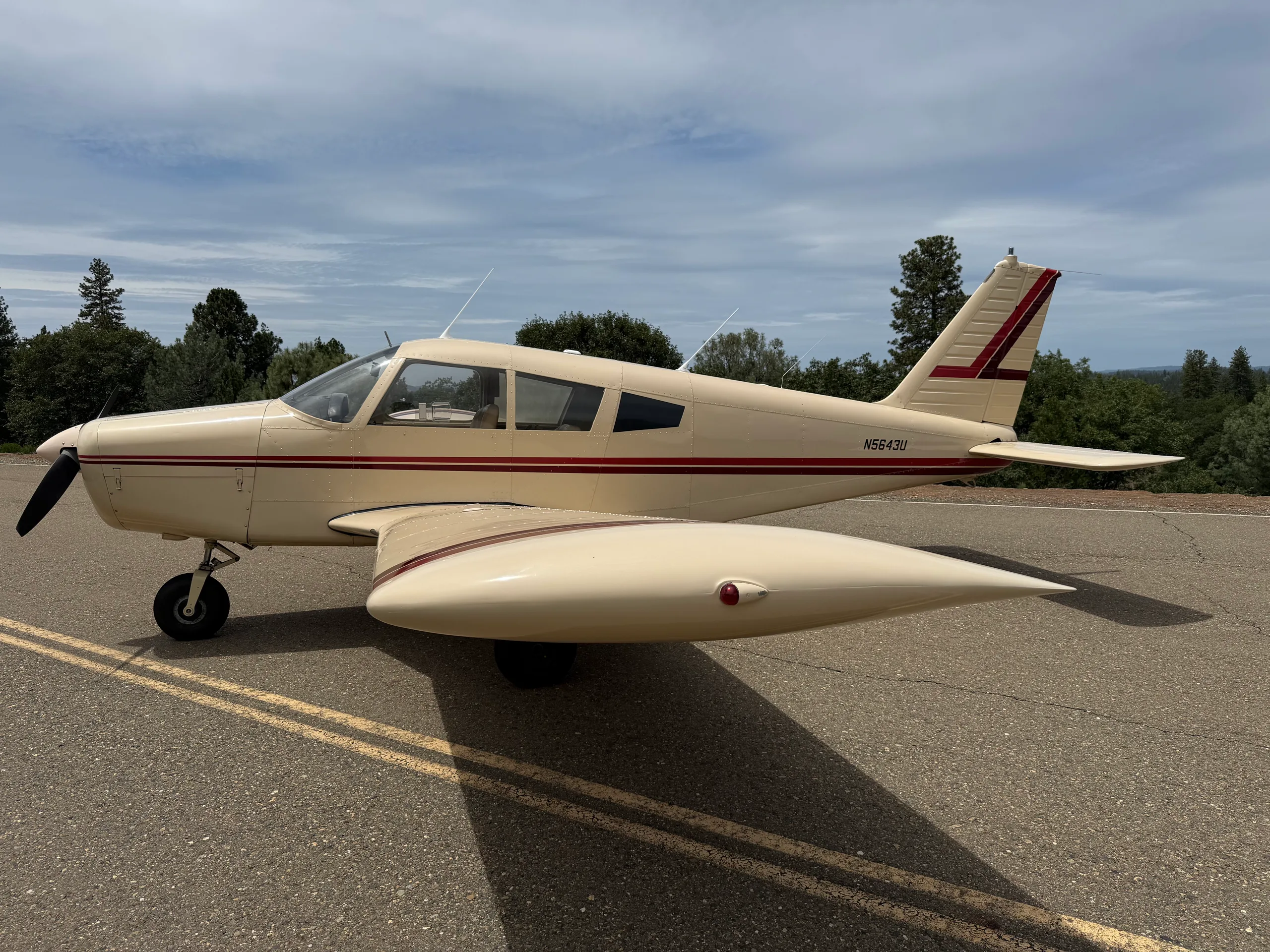 A cream-colored single-engine propeller aircraft with red stripes on the fuselage and tail, registration N5643U, parked on an asphalt surface with trees in the background.