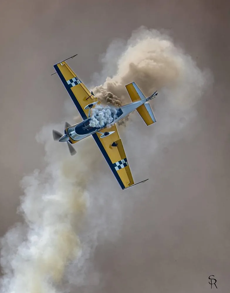 A yellow aerobatic aircraft with checkerboard wings performs a maneuver, surrounded by smoke.