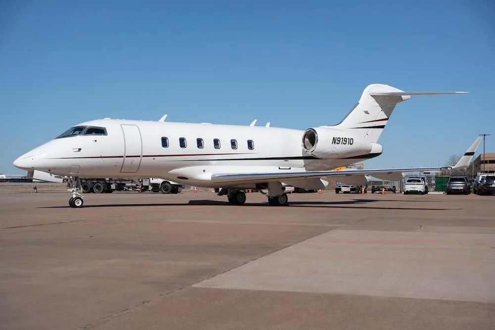 A white Bombardier Challenger business jet with registration N91910 is parked on an airport tarmac under a clear blue sky.