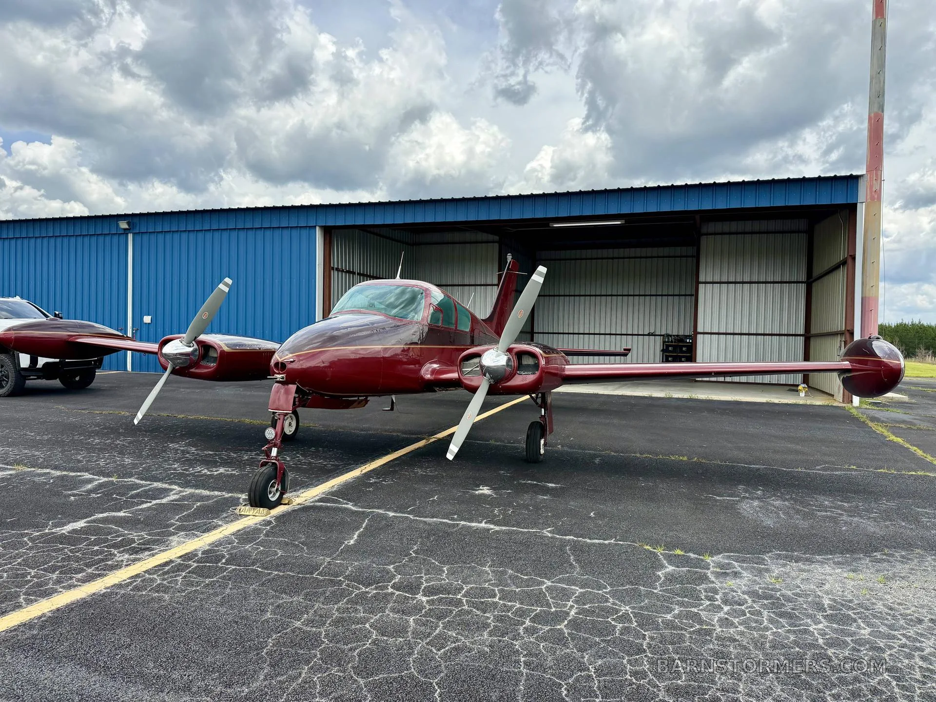 A front view of a red twin-engine aircraft, likely a Beechcraft Baron, with registration N6792T, parked on an asphalt apron in front of an open blue hangar.