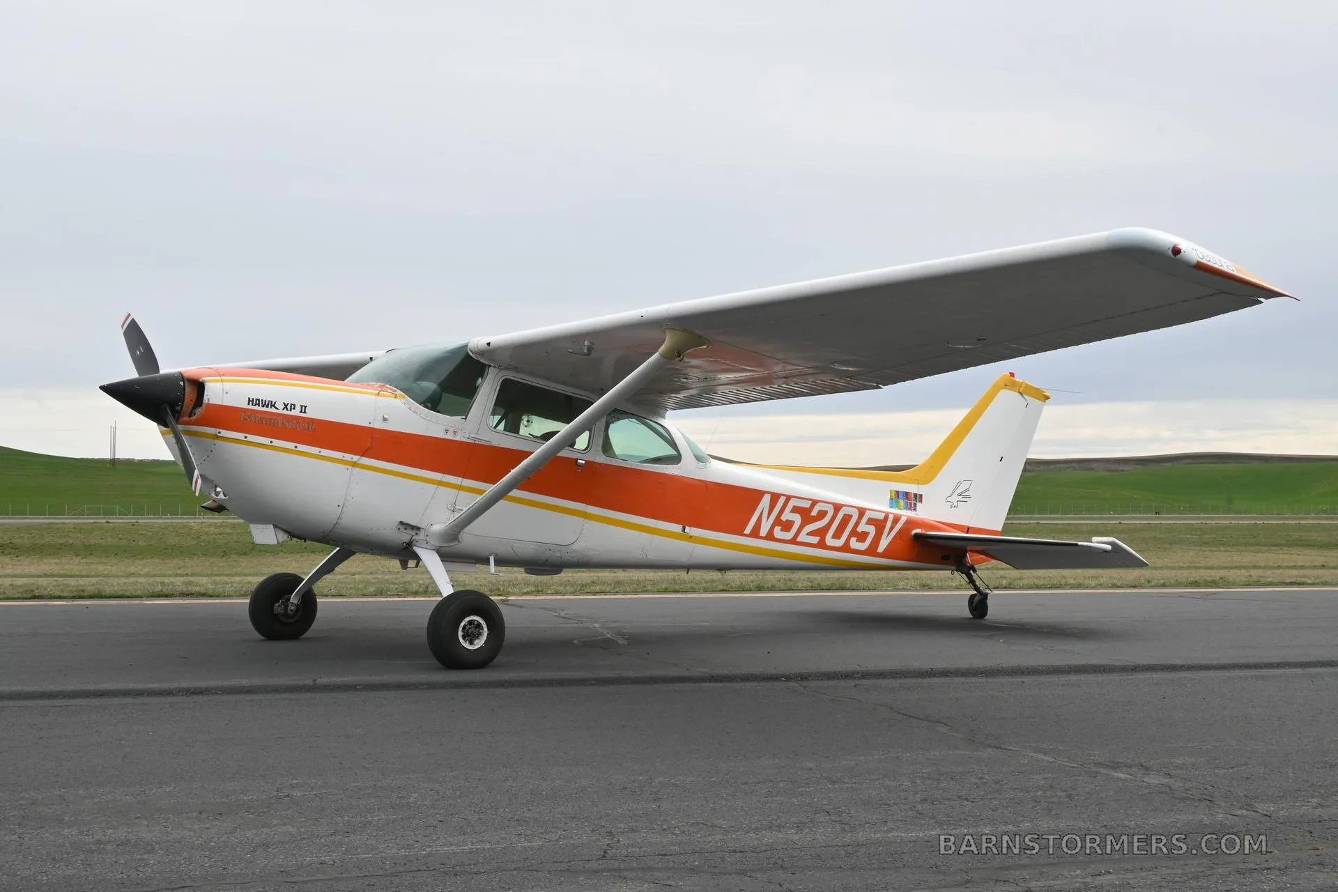 A white and orange Cessna 172 Hawk XP II (N5205V) parked on an asphalt runway with green hills in the background.