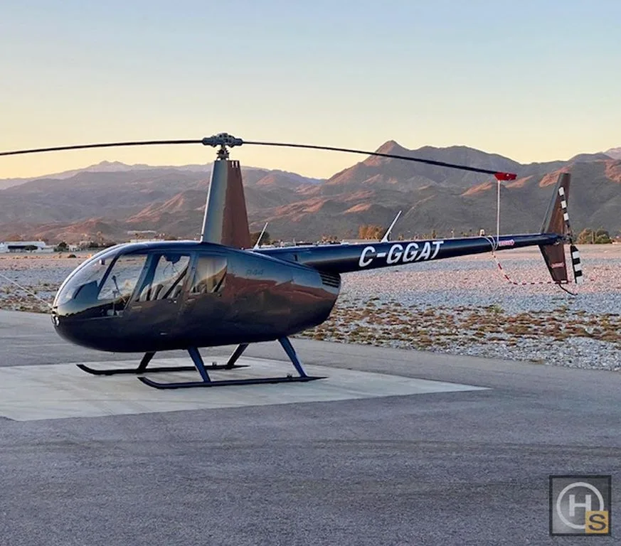 A dark blue Robinson R44 helicopter, registration C-GGAT, parked on a tarmac with mountains in the background at sunset.
