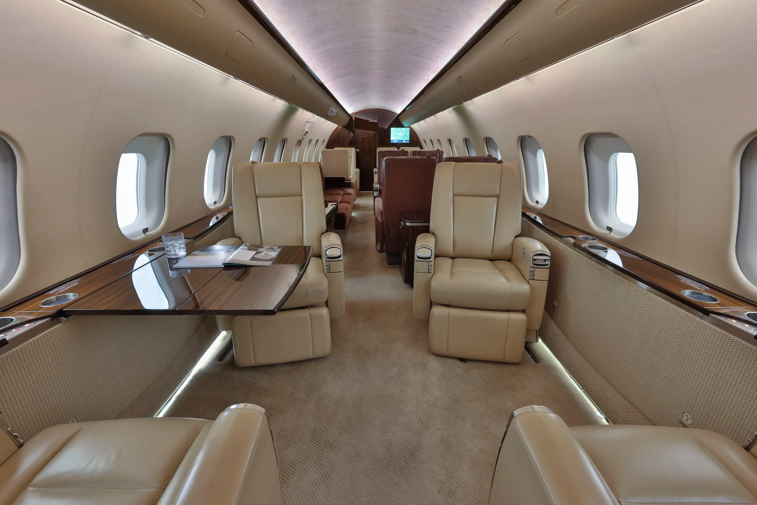 The luxurious cabin interior of a business jet, featuring beige leather seats, a polished wooden table, and large windows.