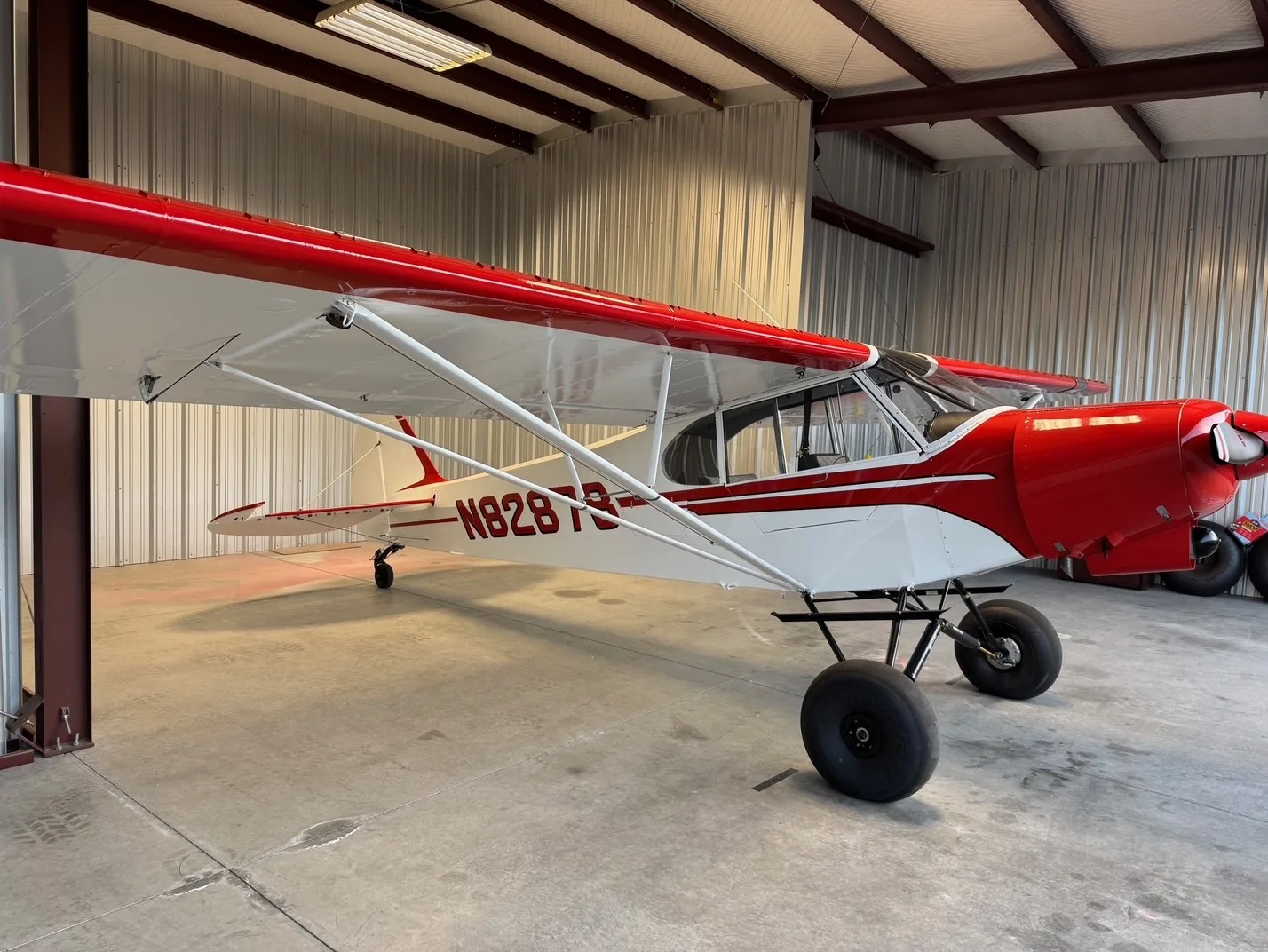 A close-up side view of the red and white bush plane, N8287B, with large tires, inside a hangar.