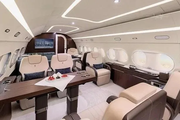 A luxurious aircraft cabin with light beige leather seats, a dark wood dining table set with fruit, and a large screen displaying a night city view.