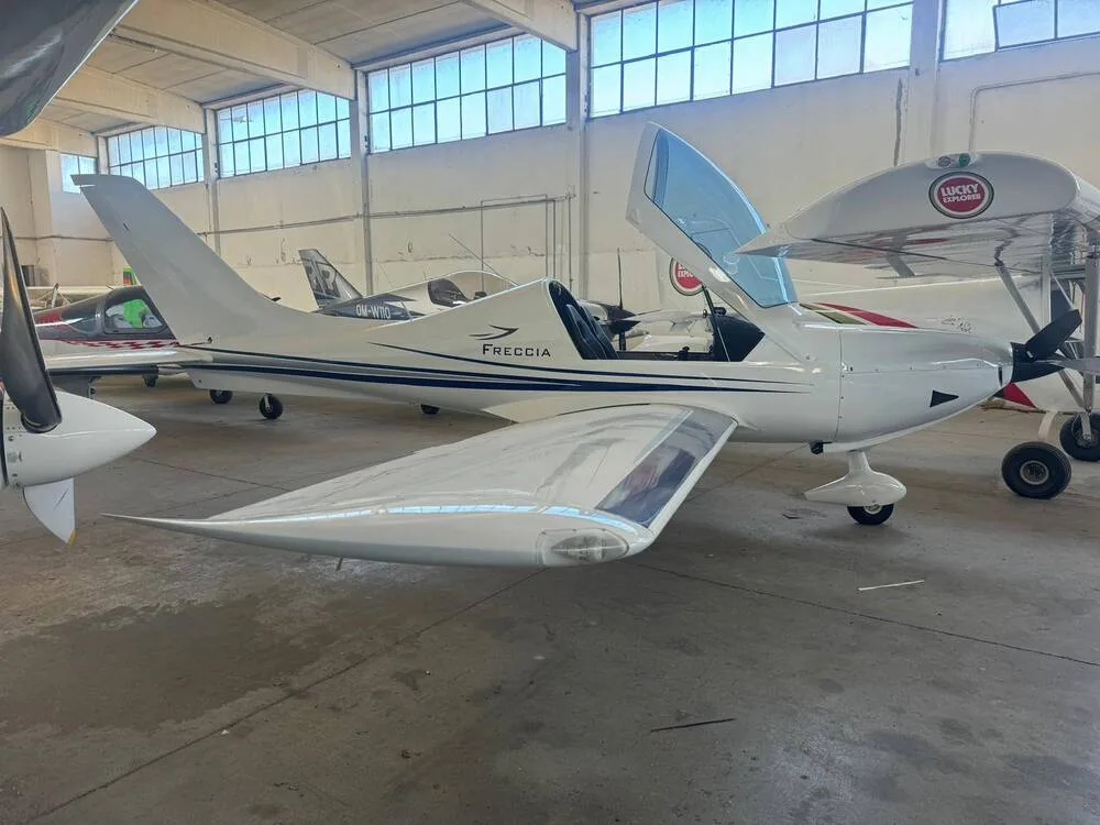 A white Freccia light aircraft with its canopy open, parked in a hangar next to other planes.