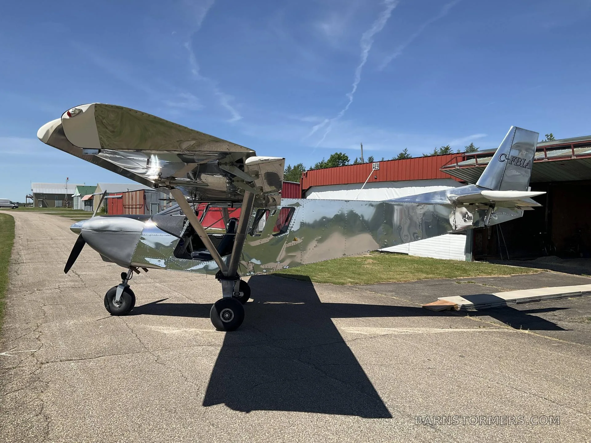 A shiny silver aircraft with registration C-FBIA is parked on an asphalt pad in front of a red hangar under a clear blue sky.