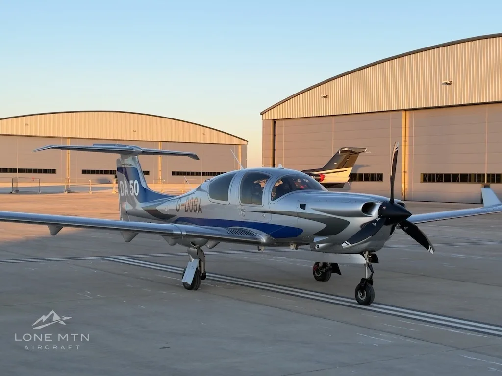 A silver and blue Diamond DA50 aircraft with registration G-DOGA parked on an airfield in front of hangars at sunset.