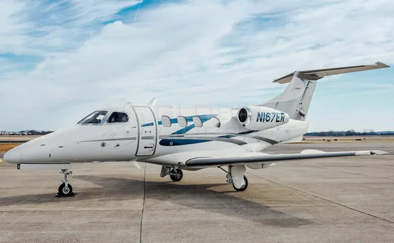 A white Embraer Phenom 300 business jet with blue and grey stripes on the fuselage, parked on an airfield under a partly cloudy sky.