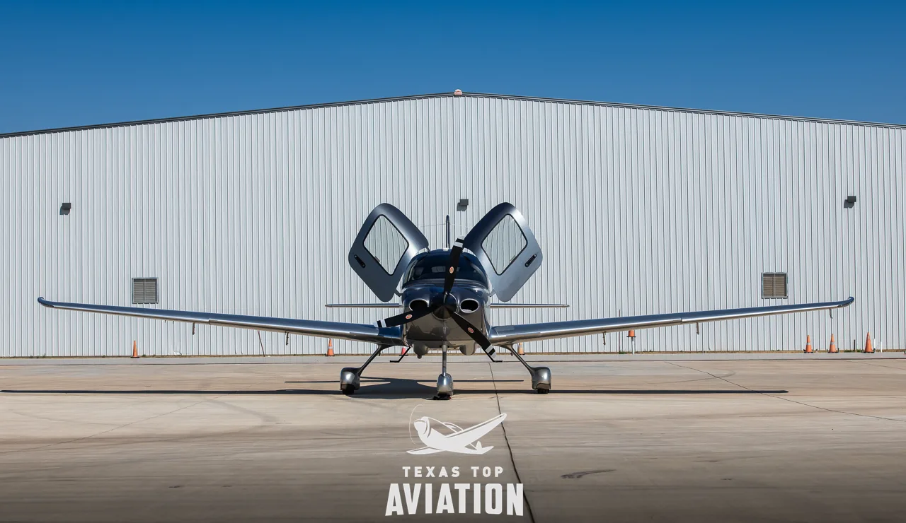A front view of a silver and grey Cirrus SR22 aircraft with registration N886CB and open doors, parked in front of a hangar.