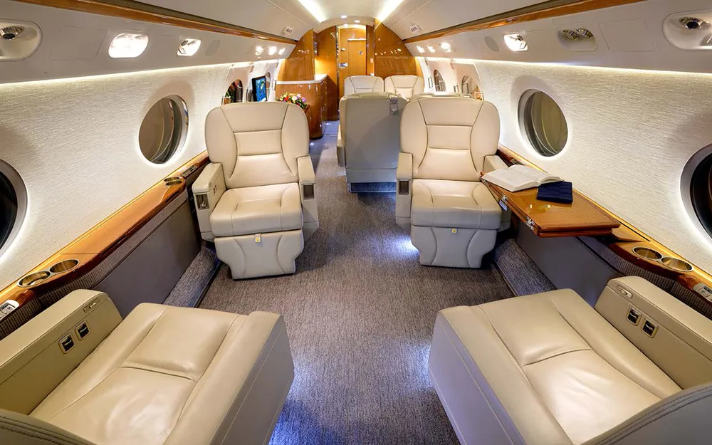 The spacious aircraft cabin with multiple beige leather seats, plush carpeting, and a book resting on a side table.