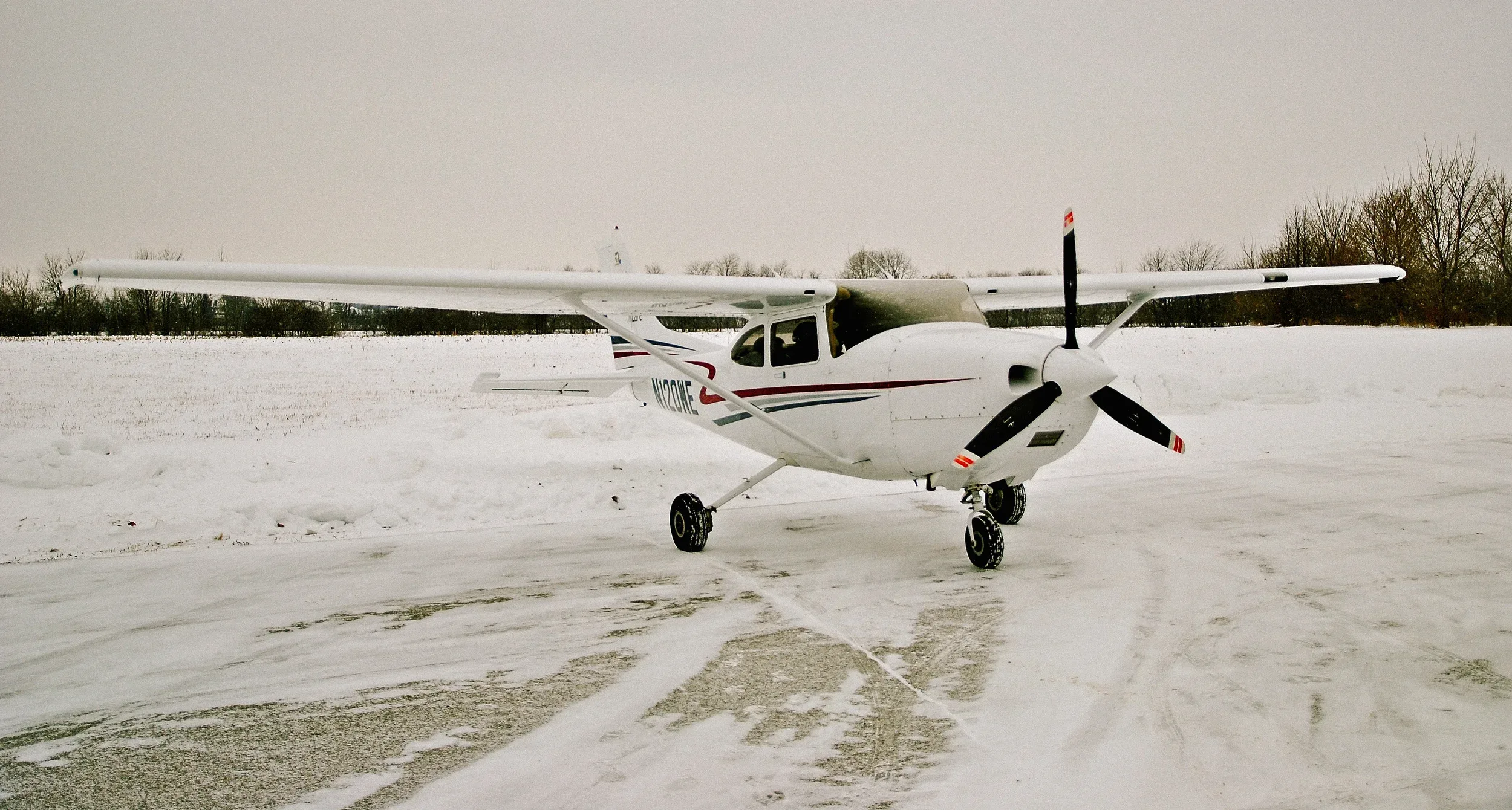 A white single-engine high-wing aircraft with red and blue stripes is parked on a snow-covered field under an overcast sky.