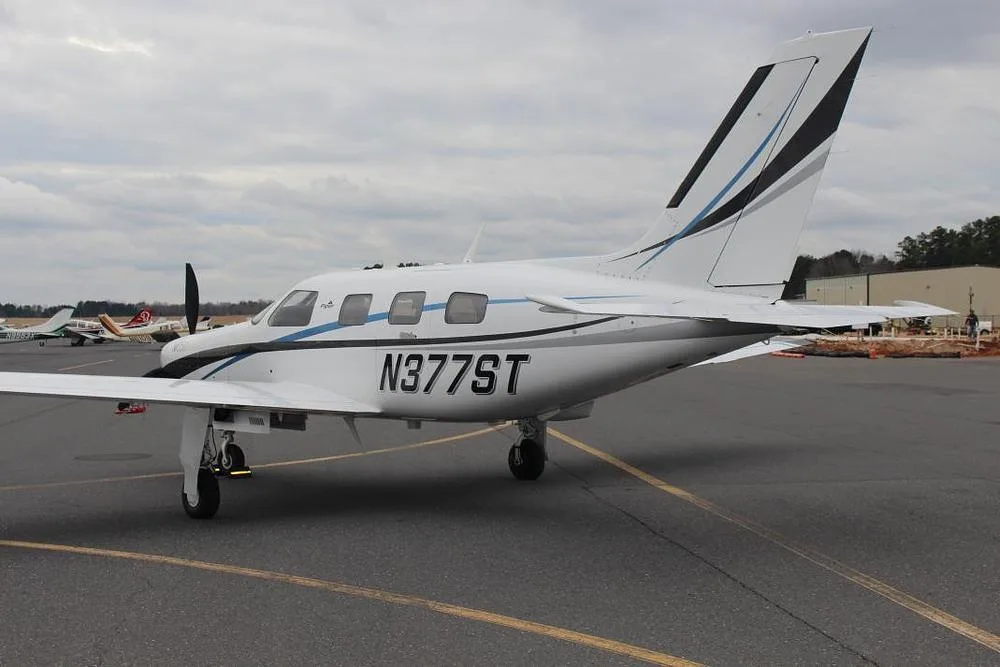 A rear-side view of a white Piper M350 aircraft with black and blue stripes, registration N377ST, parked on an asphalt tarmac.
