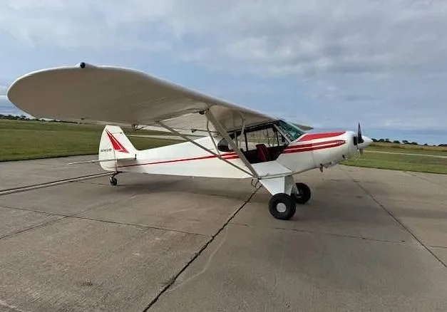 A white and red single-engine taildragger aircraft is parked on a concrete tarmac, viewed from the side.