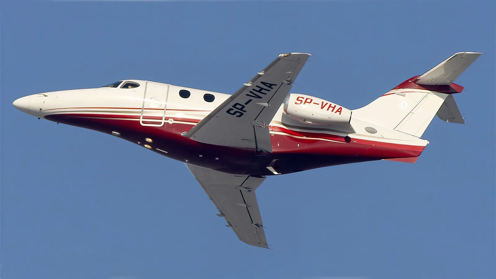 A white and red business jet, tail number SP-VHA, is seen flying against a clear blue sky.