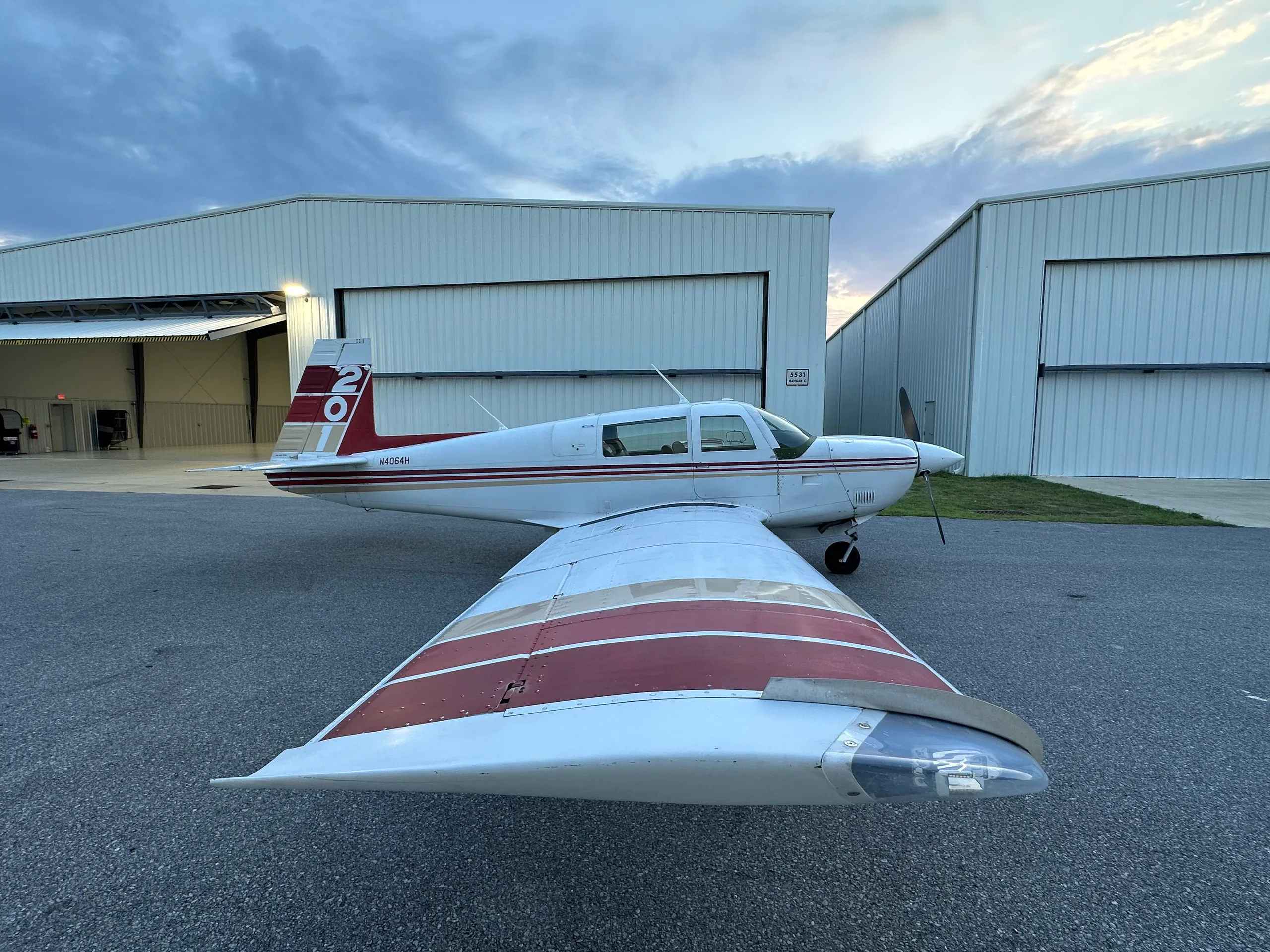 A white single-engine aircraft with red and gold stripes on the tail and wings, parked on an asphalt apron outside hangars at dusk.