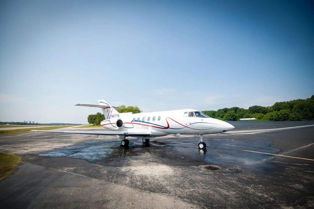 A white business jet with red and blue stripes (N8778) is parked on a wet tarmac with trees and distant buildings.
