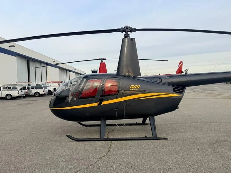 A black Robinson R44 Raven II helicopter with a yellow stripe, parked on an airfield next to a hangar.