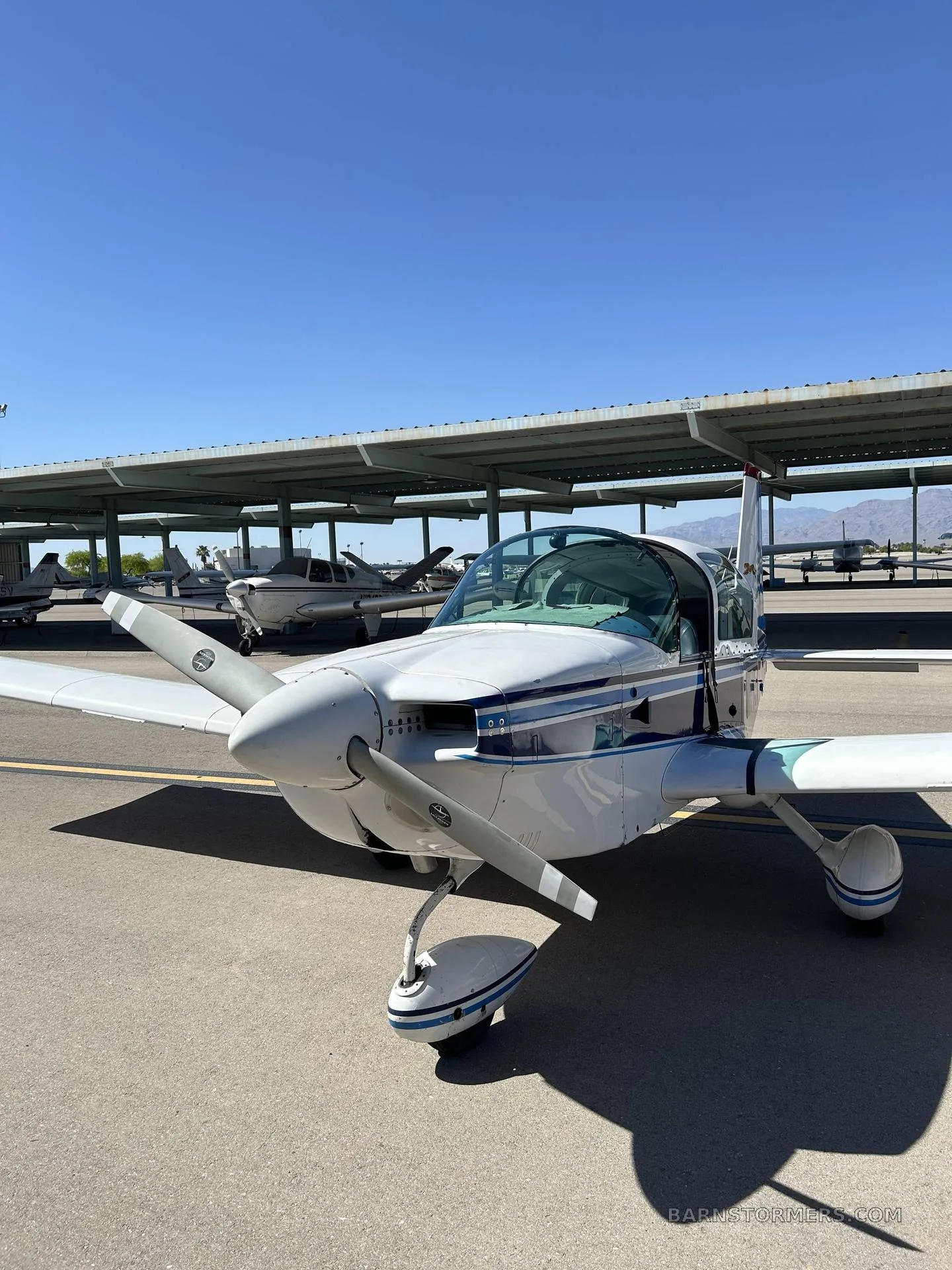 A close-up front view of a white and blue Piper Cherokee aircraft (N9869U) parked on an asphalt tarmac, with hangars in the background.