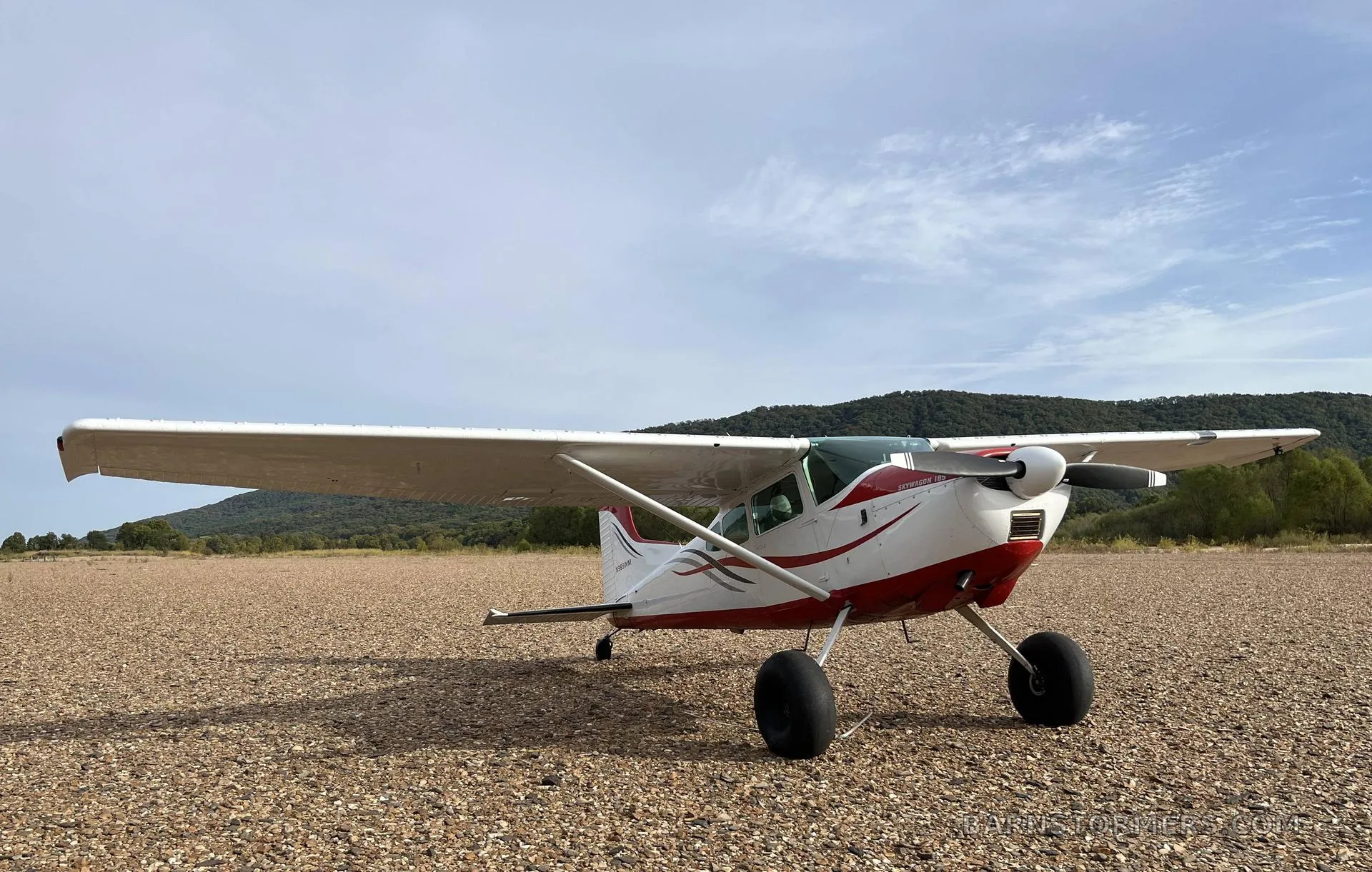 A white and red Cessna 180 aircraft with tundra tires parked on a wide gravel area under a partly cloudy sky.