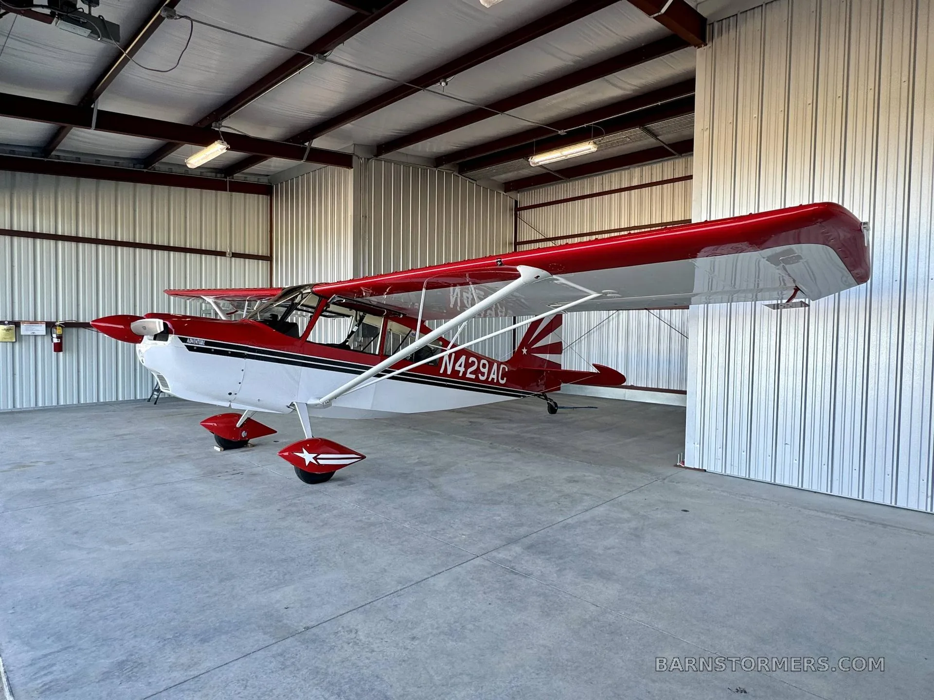 A red and white Adventure aircraft, N429AC, with a star on the wheel fairings, parked inside a hangar.