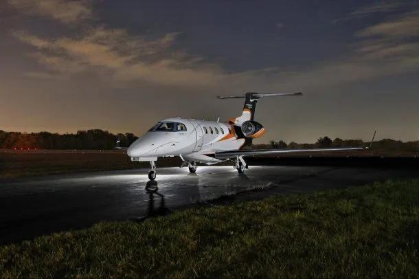 A white private jet with orange and black accents sits on a wet tarmac at night, illuminated by its landing lights.