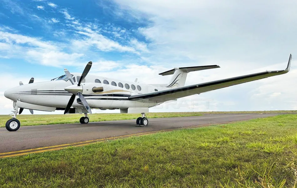 A white Beechcraft King Air twin-turboprop aircraft with black and gold stripes is parked on a runway under a partly cloudy sky.