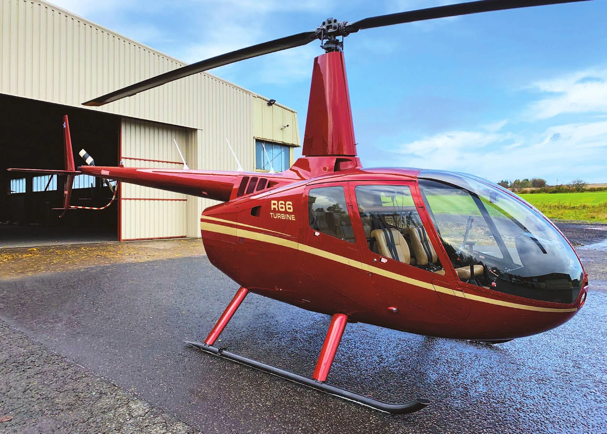 A red Robinson R66 Turbine helicopter with gold stripes parked on a wet tarmac next to a hangar.