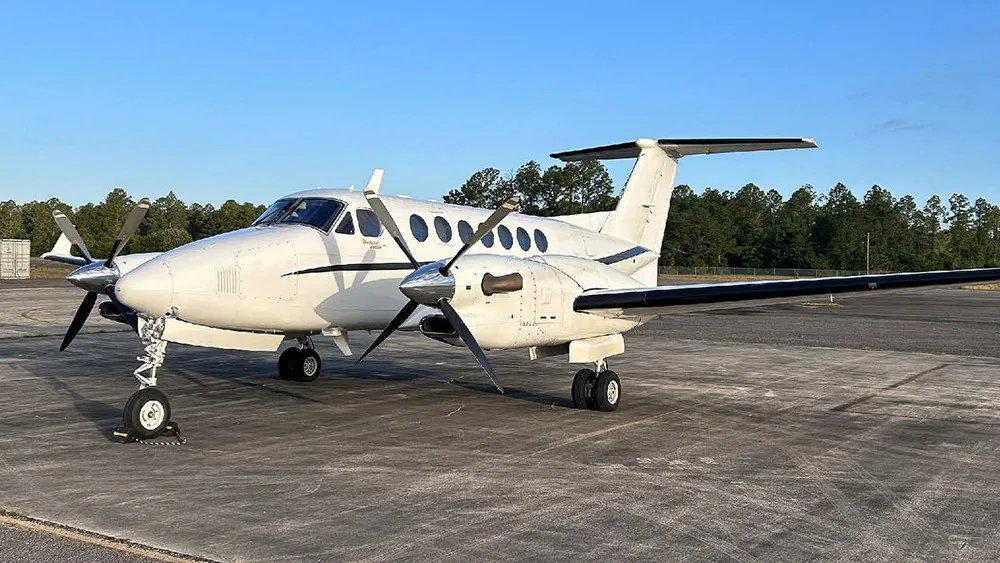 A white Beechcraft King Air 350 turboprop aircraft is parked on an asphalt tarmac with trees in the background.