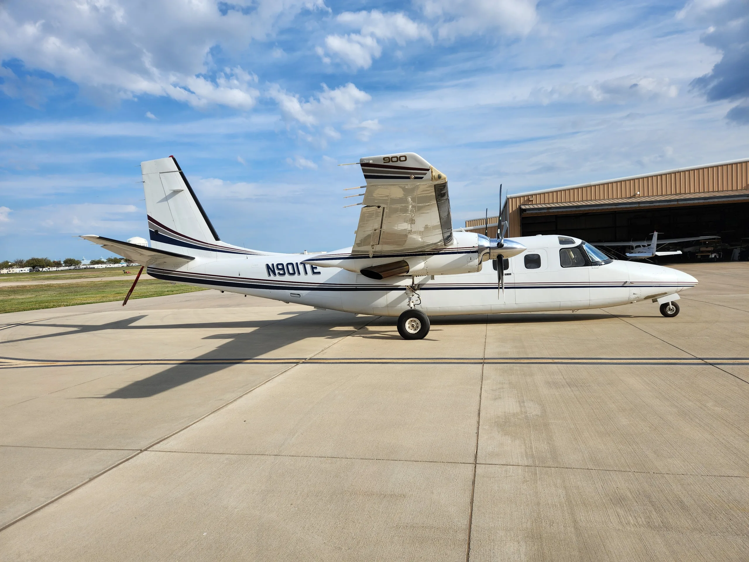 A white twin-engine propeller aircraft with dark blue and gold stripes, registration N901TE, parked on a concrete apron with a hangar in the background.