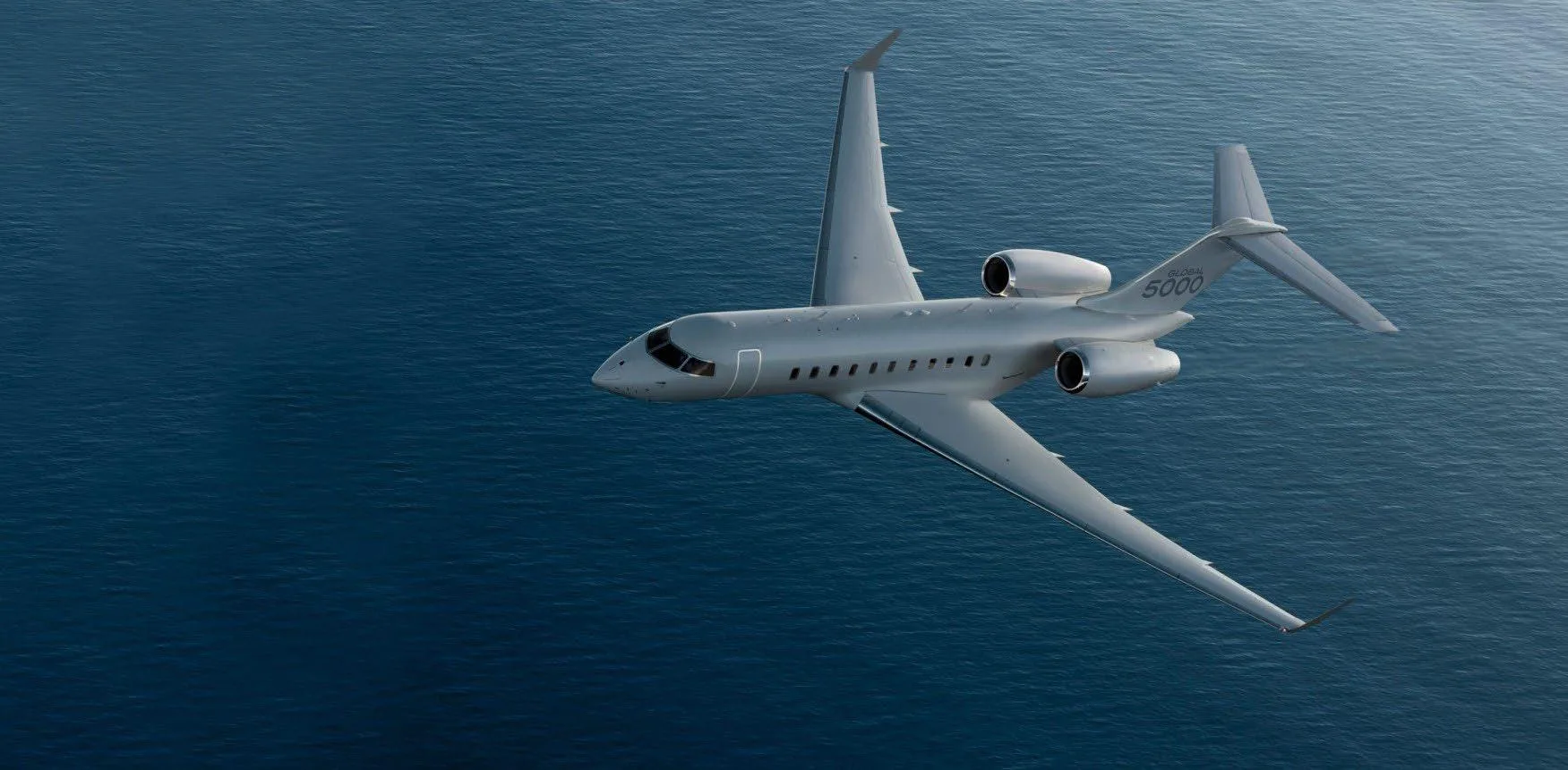 A grey Bombardier Global 5000 business jet flies over a blue ocean.