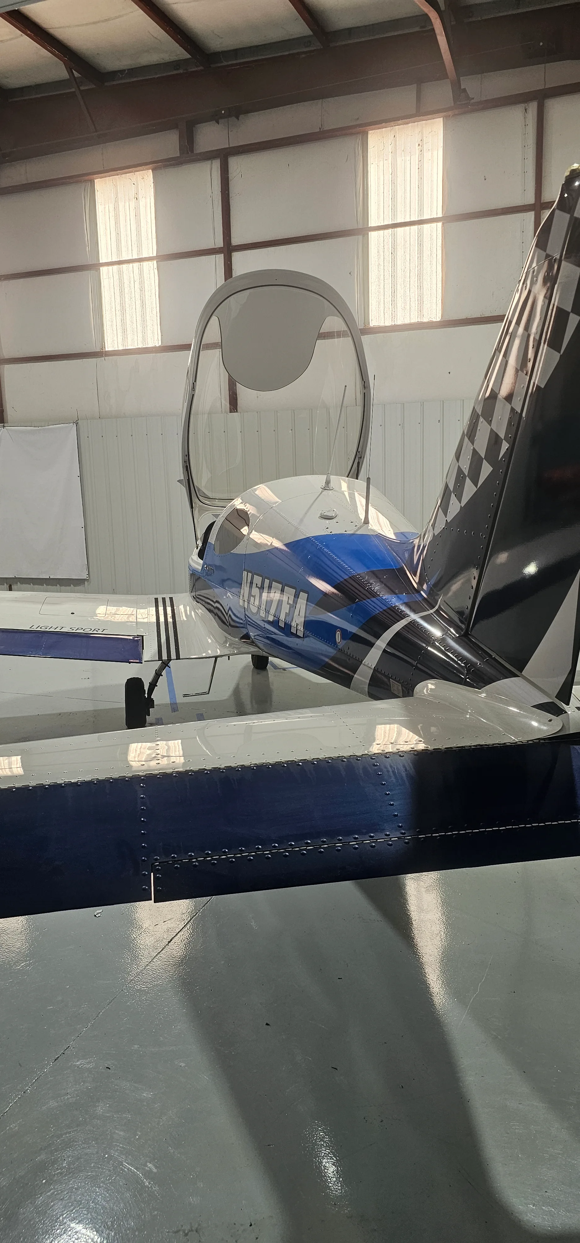 A side view of a white and blue light sport aircraft with an open canopy, parked inside a hangar.