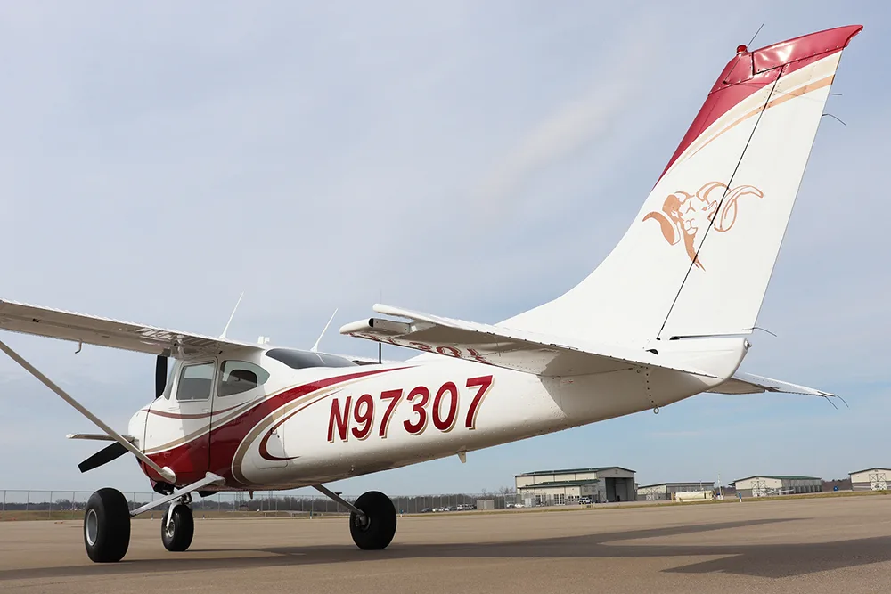 A rear-side view of a white and red single-engine high-wing aircraft, N97307, parked on an airfield with hangars in the background.