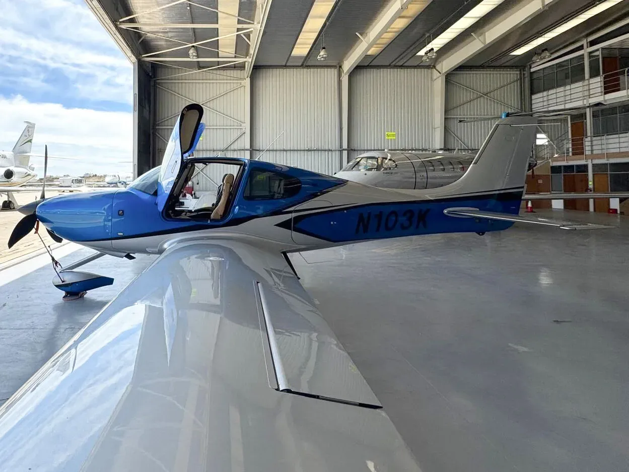 A blue and silver Cirrus SR22 aircraft (N103K) with open doors, viewed from the right side in a hangar.