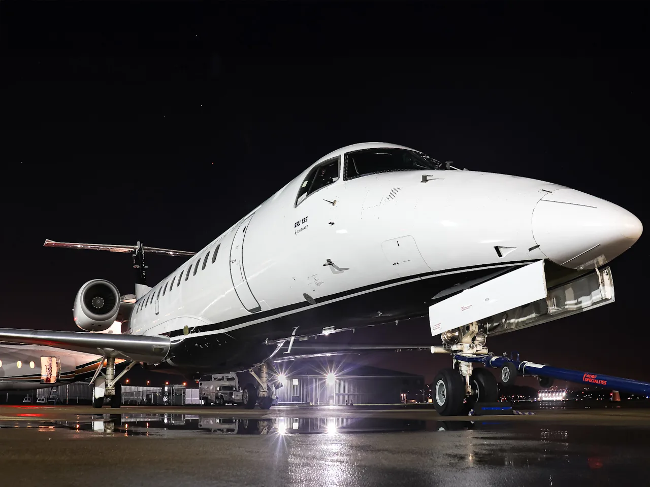A white and black Embraer ERJ 135 aircraft is parked on a wet tarmac at night, illuminated by ground lights.