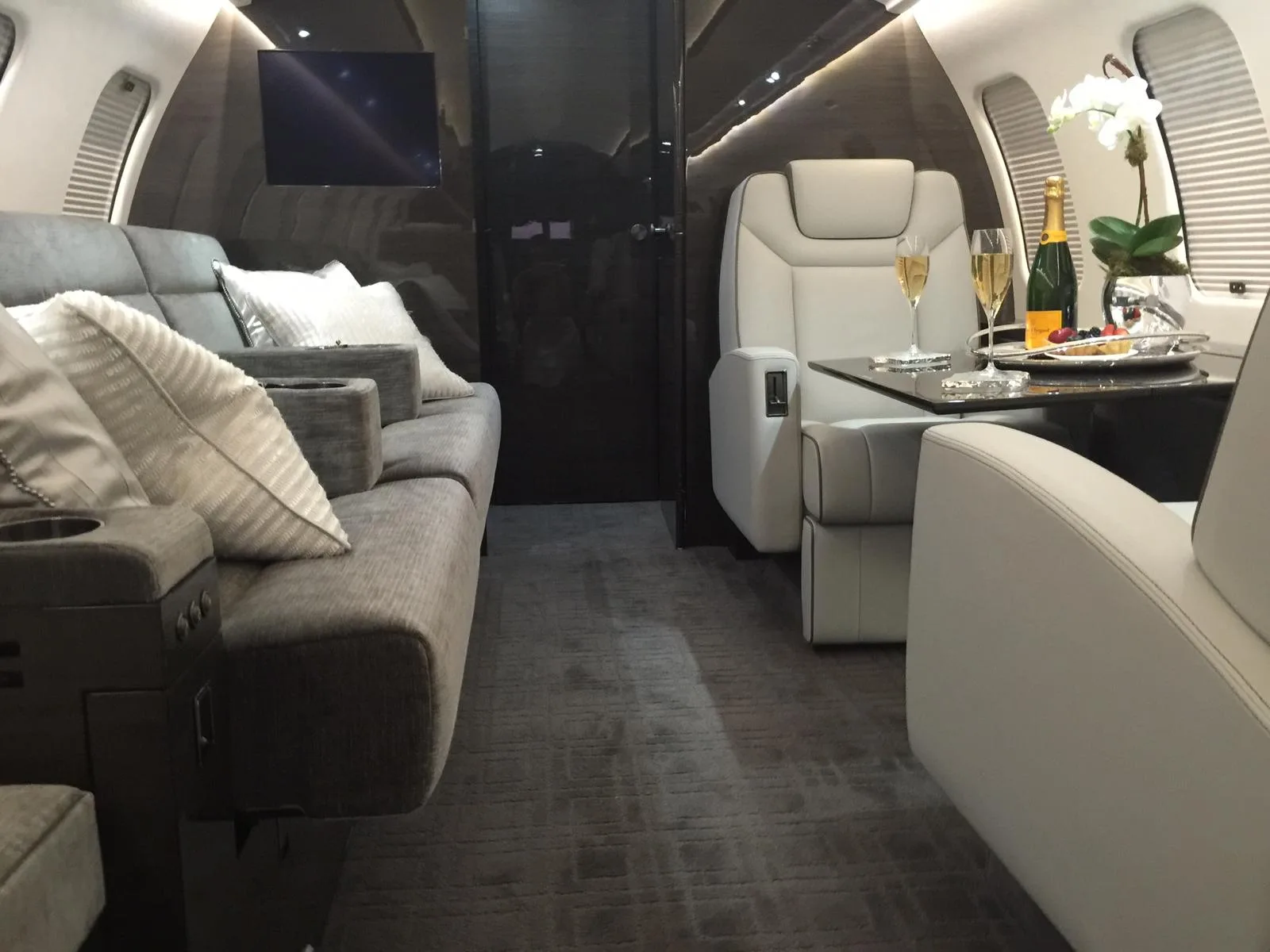 The luxurious interior cabin of a private jet, featuring grey sofas, a white armchair, and a table set with champagne and fruit.