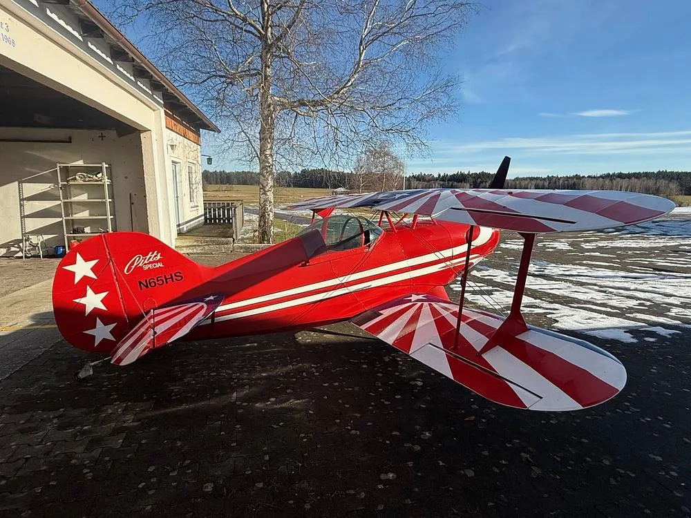A red Pitts Special biplane with white and red striped wings and tail, parked on an asphalt surface with a snowy field in the background.