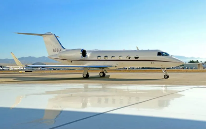 A white Gulfstream G550 business jet with a blue tail stripe is parked on an airport tarmac under a clear blue sky.