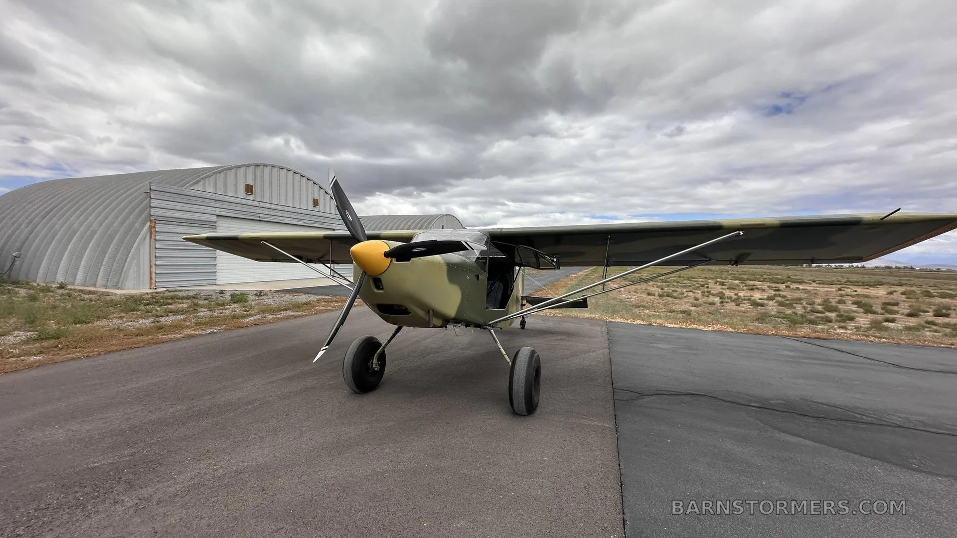 A light aircraft with camouflage paint and a yellow propeller spinner is parked on an asphalt runway in front of hangars under a cloudy sky.