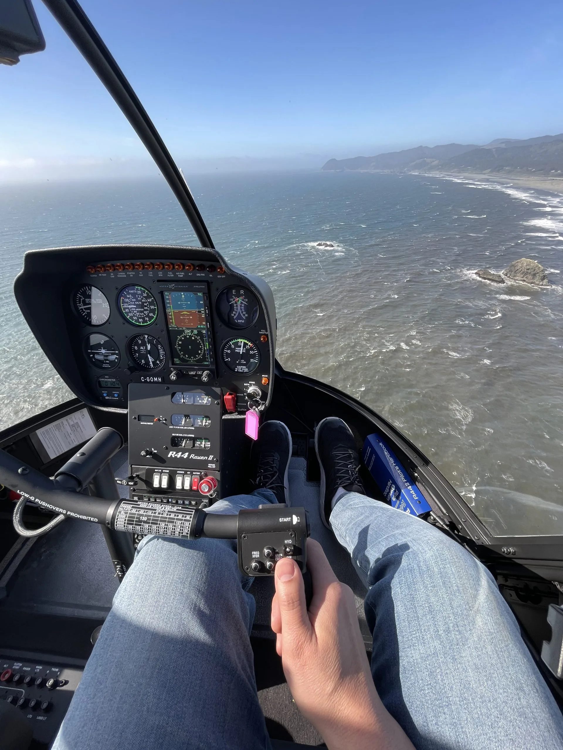 A pilot's view from the cockpit of a Robinson R44 Raven II helicopter, showing the instrument panel and a scenic coastline over the ocean.