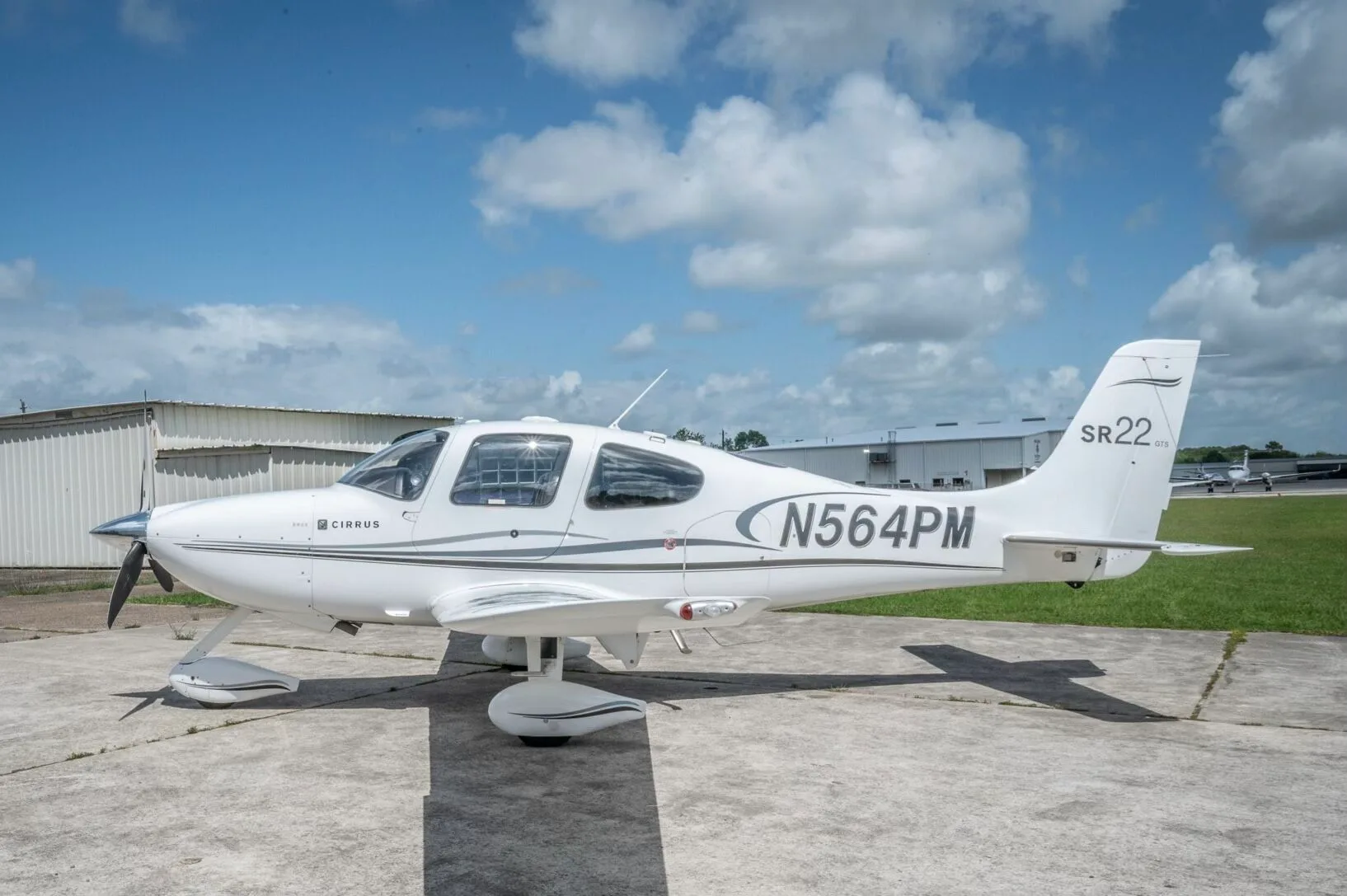 A white Cirrus SR22 GTS aircraft with N564PM registration parked on a concrete ramp under a partly cloudy sky.
