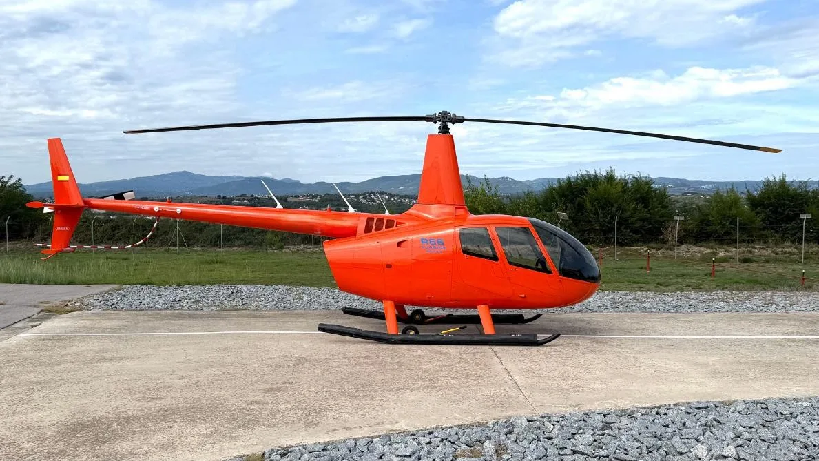 A bright orange Robinson R66 helicopter parked on a concrete pad with mountains in the background.